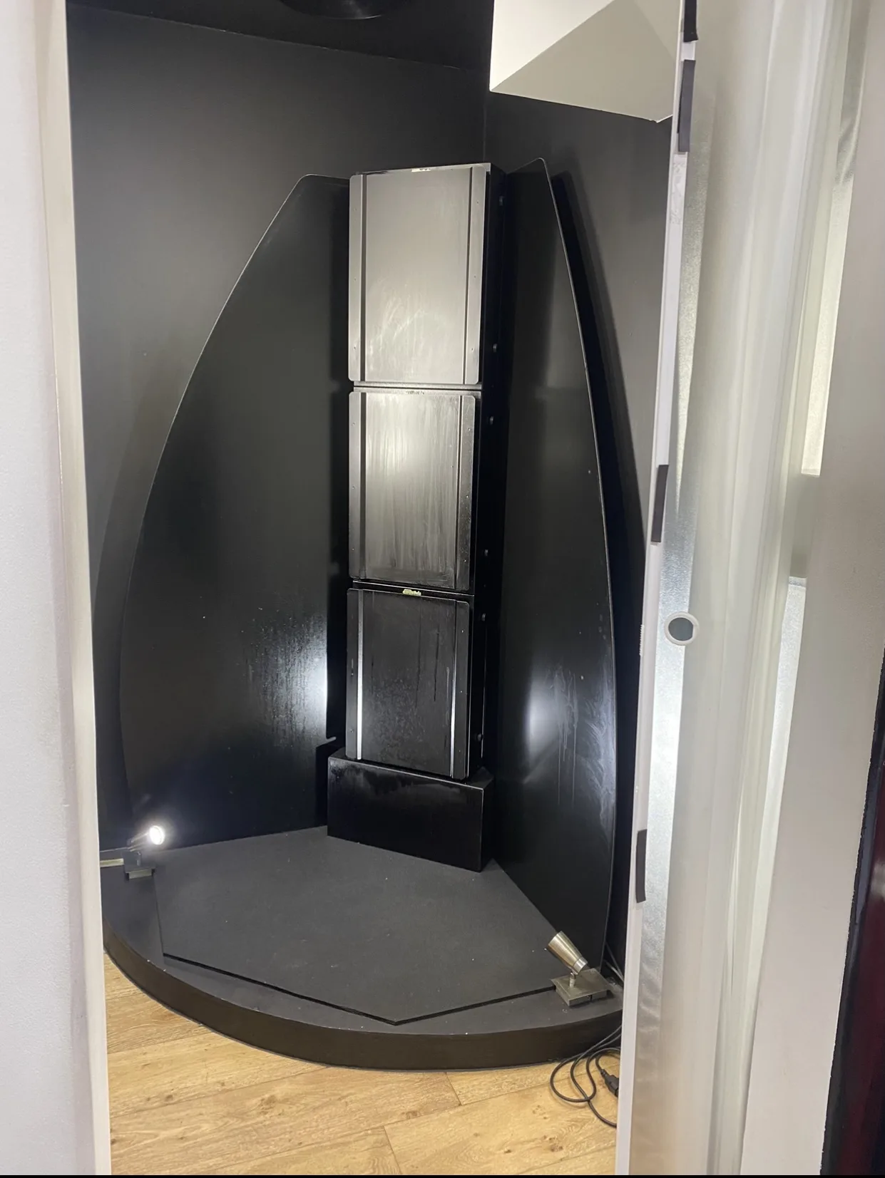 Inside Beauty B — the stand-up spray-tan booth seen through the open doorway from the treatment room, gloss-black panels and silver spray column, warm oak floorboards in the foreground