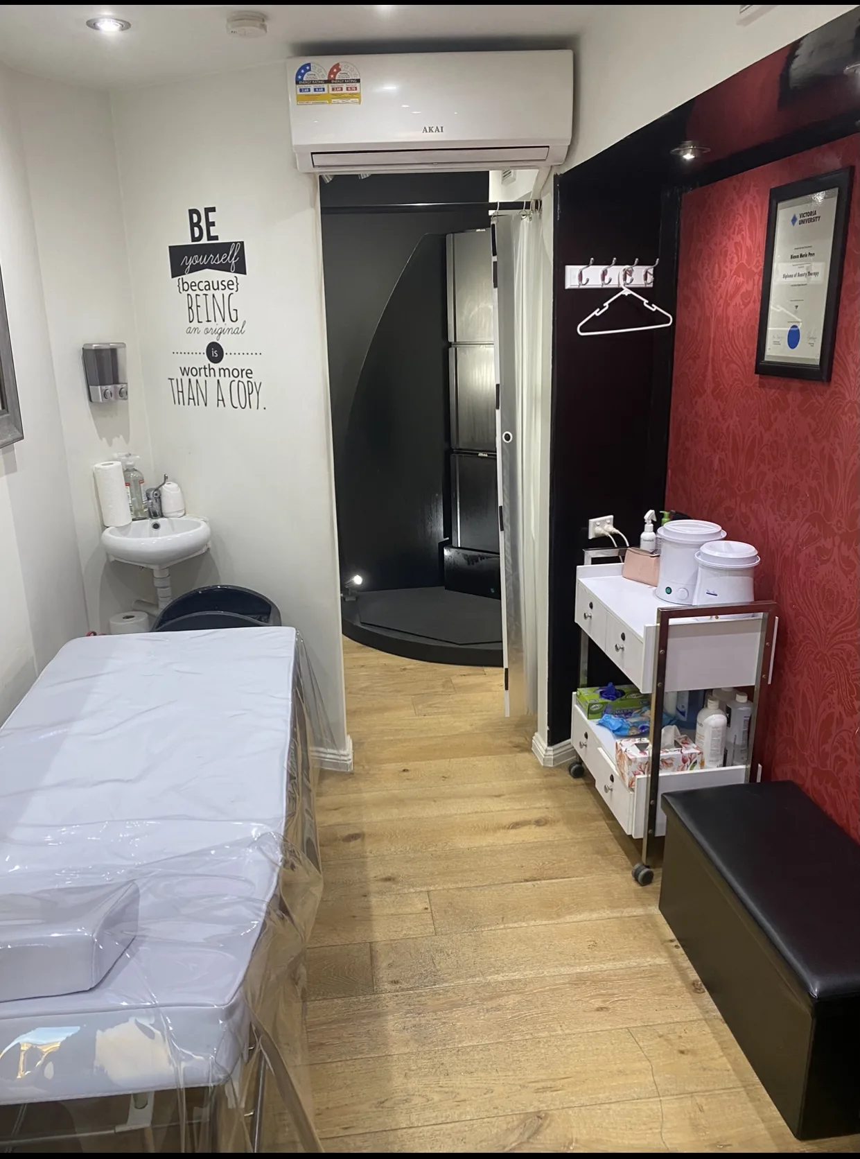 The treatment room at Beauty B — a single covered bed, wax trolley against the crimson damask wall, corner hand basin, and the spray-tan booth visible through the doorway at the far end