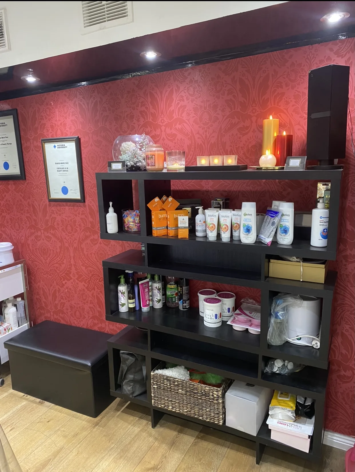 The retail shelf at Beauty B — a tall black stepped unit stocked with skincare and aftercare product against the full-wall crimson damask wallpaper, with framed Victoria University certificates to the left and lit candles along the top