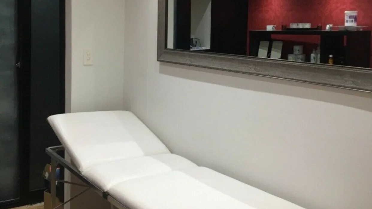 The treatment bed at Beauty B set with white linens, beneath a large mirror that reflects the red damask retail wall opposite